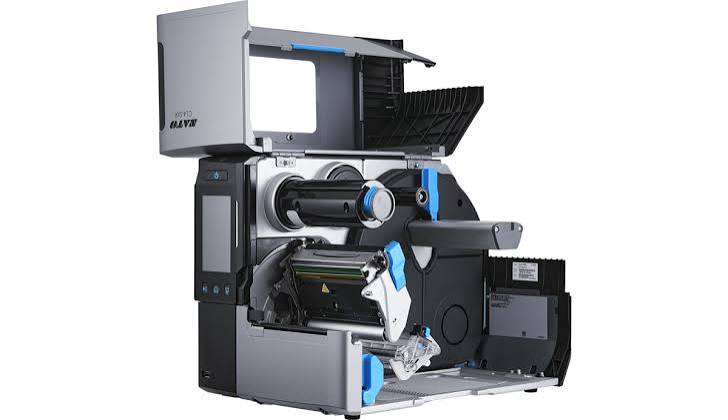 A compact industrial label printer or label application system shown in side profile on a plain white background. The machine has a dark gray and black body with bright blue accents and a partially open top cover.