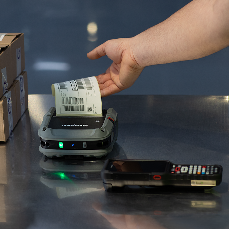 Hand placing a barcode label on a label printer with boxes and a phone in the background.