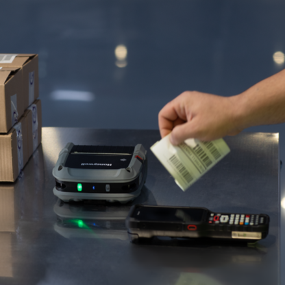 Hand holding a barcode label above a scanner on a reflective surface with cardboard boxes in the background.