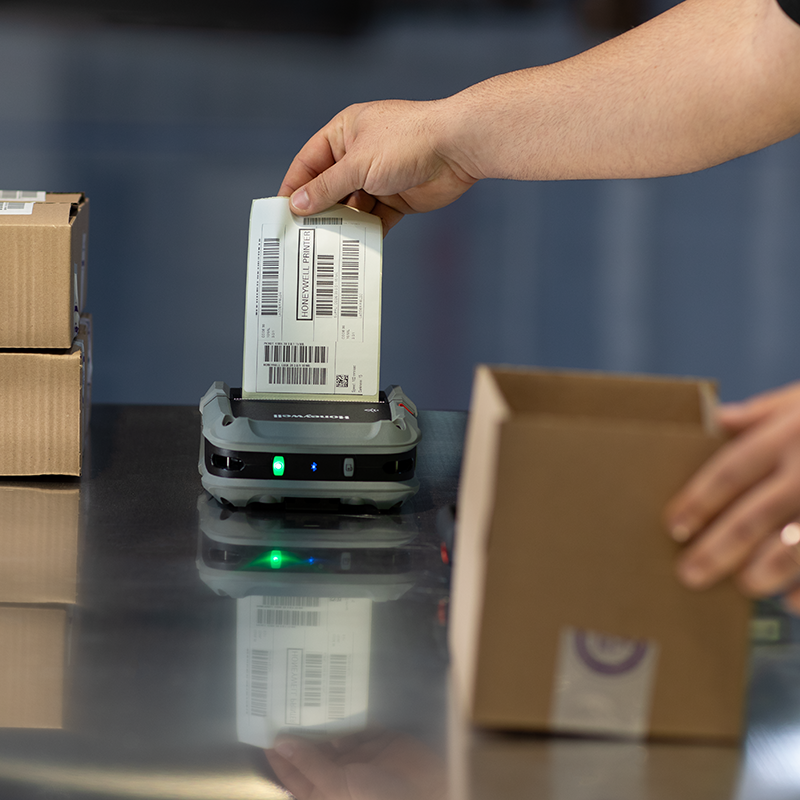 Person using a label printer to print a barcode label on a reflective surface with cardboard boxes in the background.