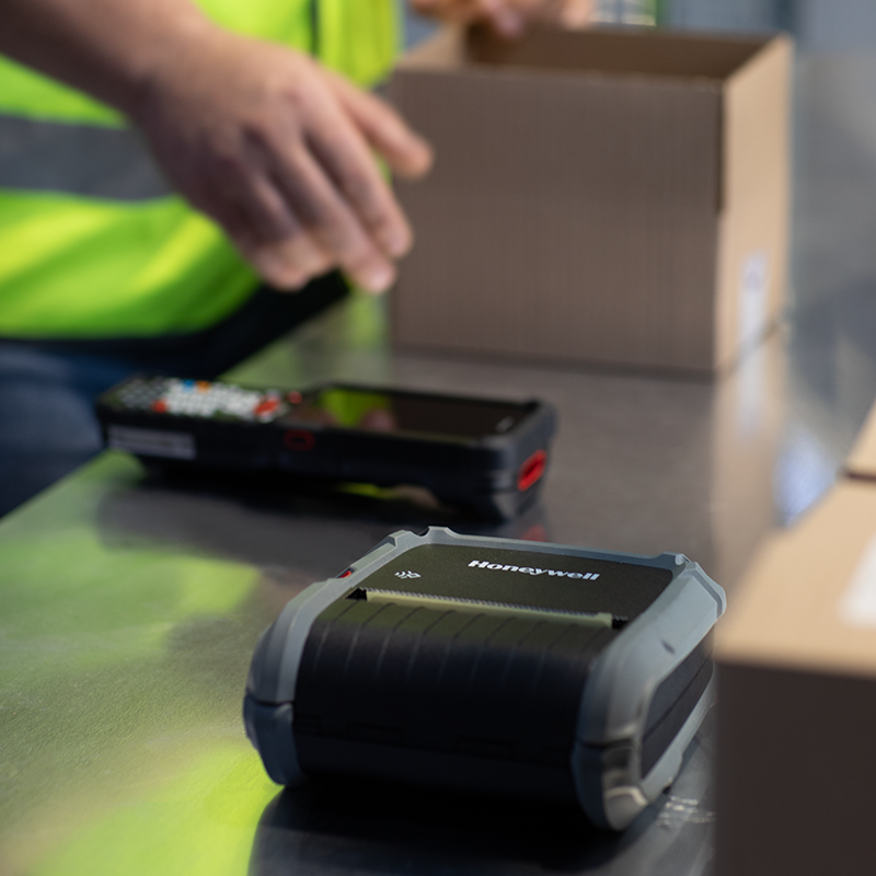 Honeywell barcode scanner on a table with a person in a high-visibility vest and a box in the background.